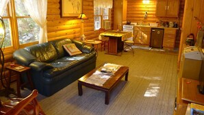 Ein gemütliches Blockhaus-Interieur mit einem Wohnzimmer, das eine Ledersofa, einen Holztisch und einen Essbereich mit einer Küche im Hintergrund zeigt.
