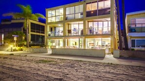 A multi-story hotel with a balcony on the second floor, a sandy beach in the foreground, and a clear night sky.