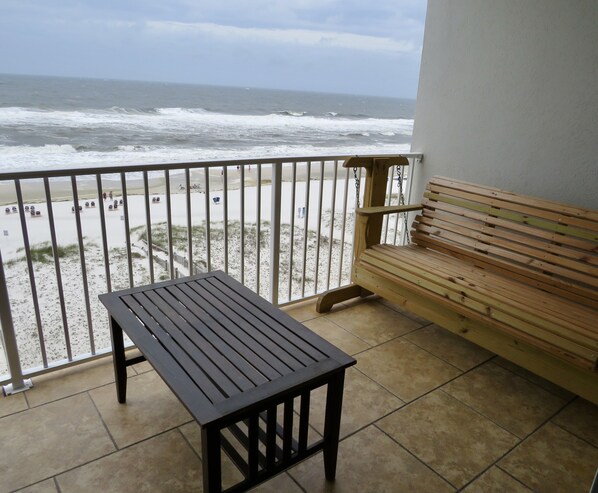 A balcony with a wooden bench and a table, overlooking a beach with people and a sandy shore. - Direct Beach Front Tropic Isles 604 (Gulf Shores)