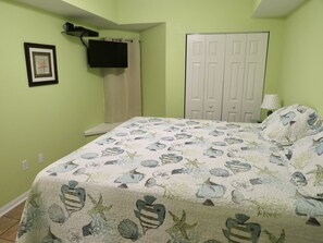 A bedroom with a bed covered in a floral-patterned quilt, a white door, and a mounted television. - Direct Beach Front Tropic Isles 604 (Gulf Shores)