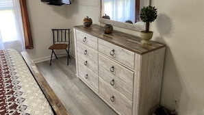 Un dormitorio con cama, cómoda, silla, televisor montado en la pared y ventana con cortinas.