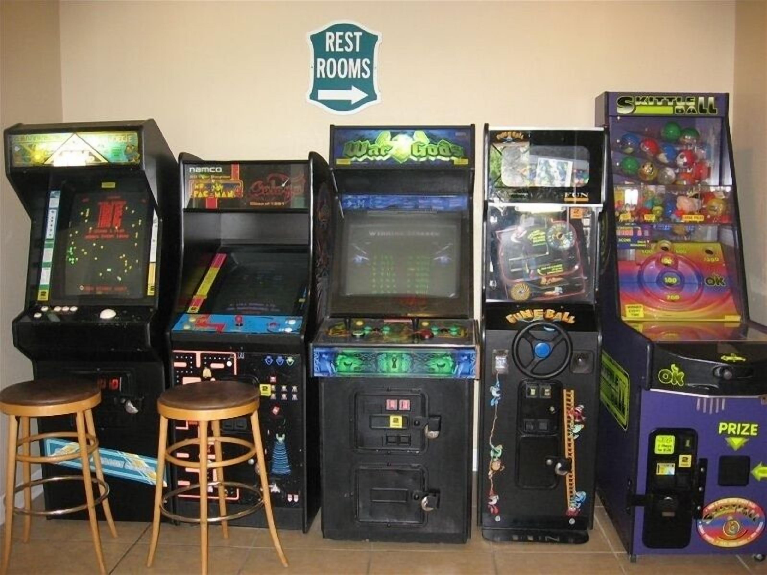 Una fila di macchinette arcade vintage con poggiapiedi di fronte, un cartello che indica i bagni e una macchina per vincere premi.