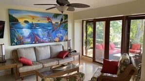 Olohuone, jossa on suuri taulu, rottingista valmistetut huonekalut ja näkymä lasiovien läpi.