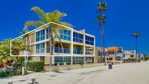 Um hotel com vários andares, grandes janelas, uma praia arenosa em primeiro plano e palmeiras.