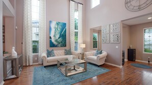 Un salón amplio con suelo de madera, una mesa de centro de cristal, dos sofás blancos con cojines azules y un cuadro abstracto de gran tamaño.