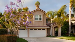 Una casa de dos pisos con garaje, una palmera y árboles con flores moradas.