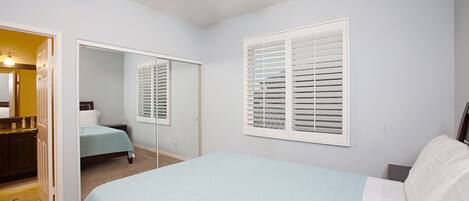 Quarto com cama, ventilador de teto, janela com persianas e espelho.