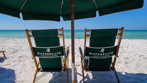 Deux fauteuils de jardin verts, portant la marque « WATERSCAPE », sont placés sur une plage de sable, avec un ciel bleu clair et l’océan en arrière-plan.
