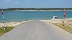 A paved pathway leading to a beach with calm blue waters, a small dock, and distant hills. - Lake Travis Retreat - Water View (Leander)
