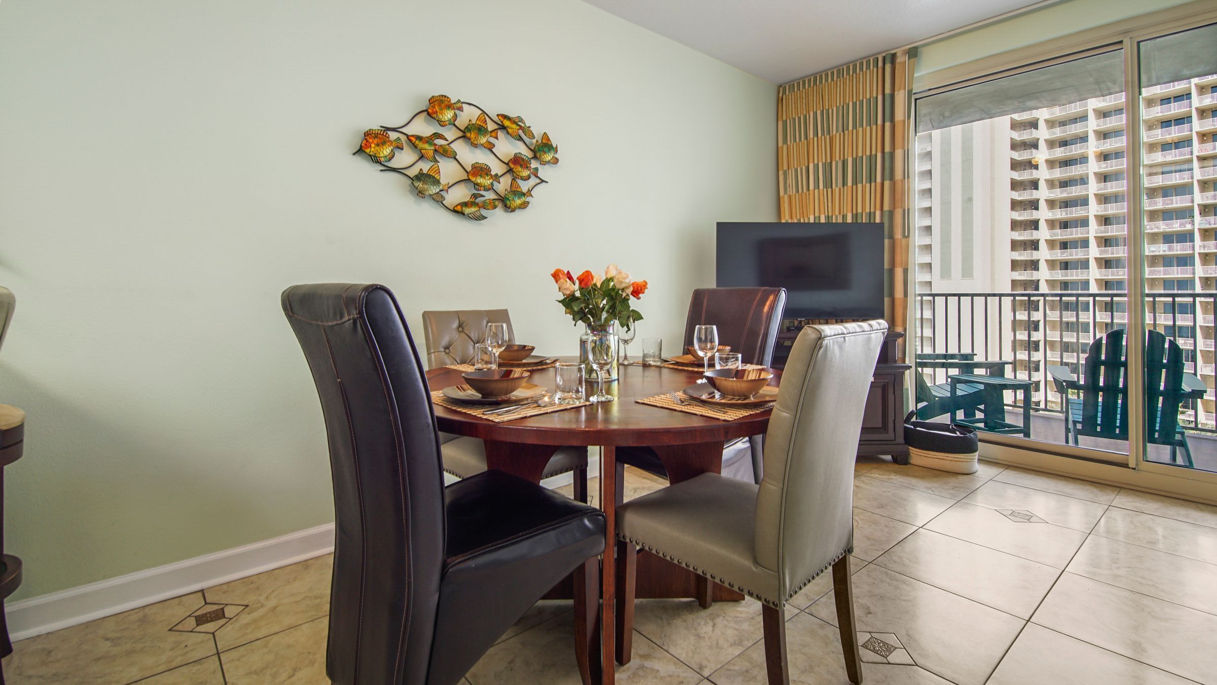 A dining area with a round table, chairs, and a vase of flowers. A flat-screen TV is mounted on the wall.