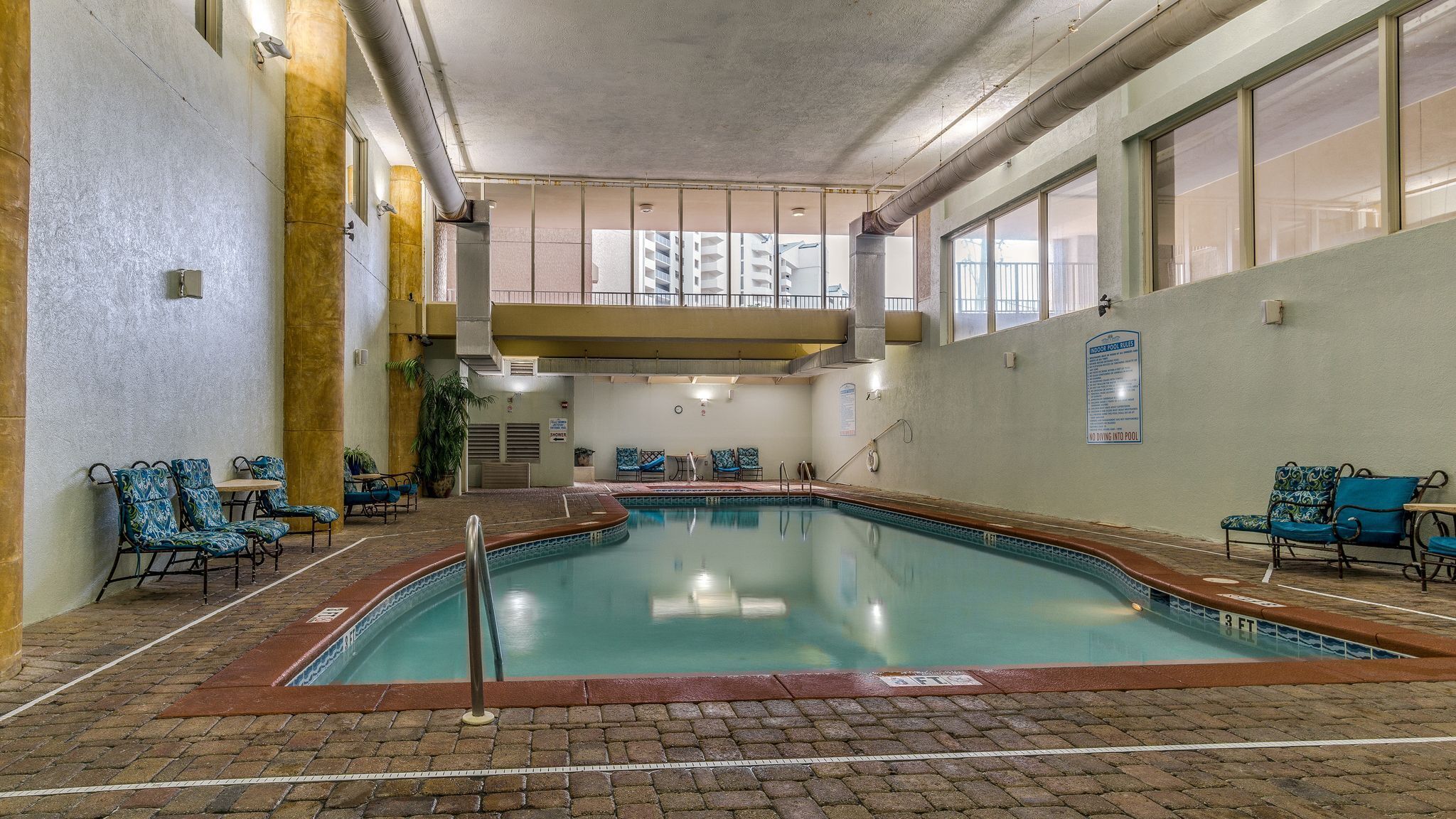 Une piscine intérieure avec un sol en briques, des chaises bleues à motifs et un panneau indiquant les règles à respecter dans la piscine intérieure.