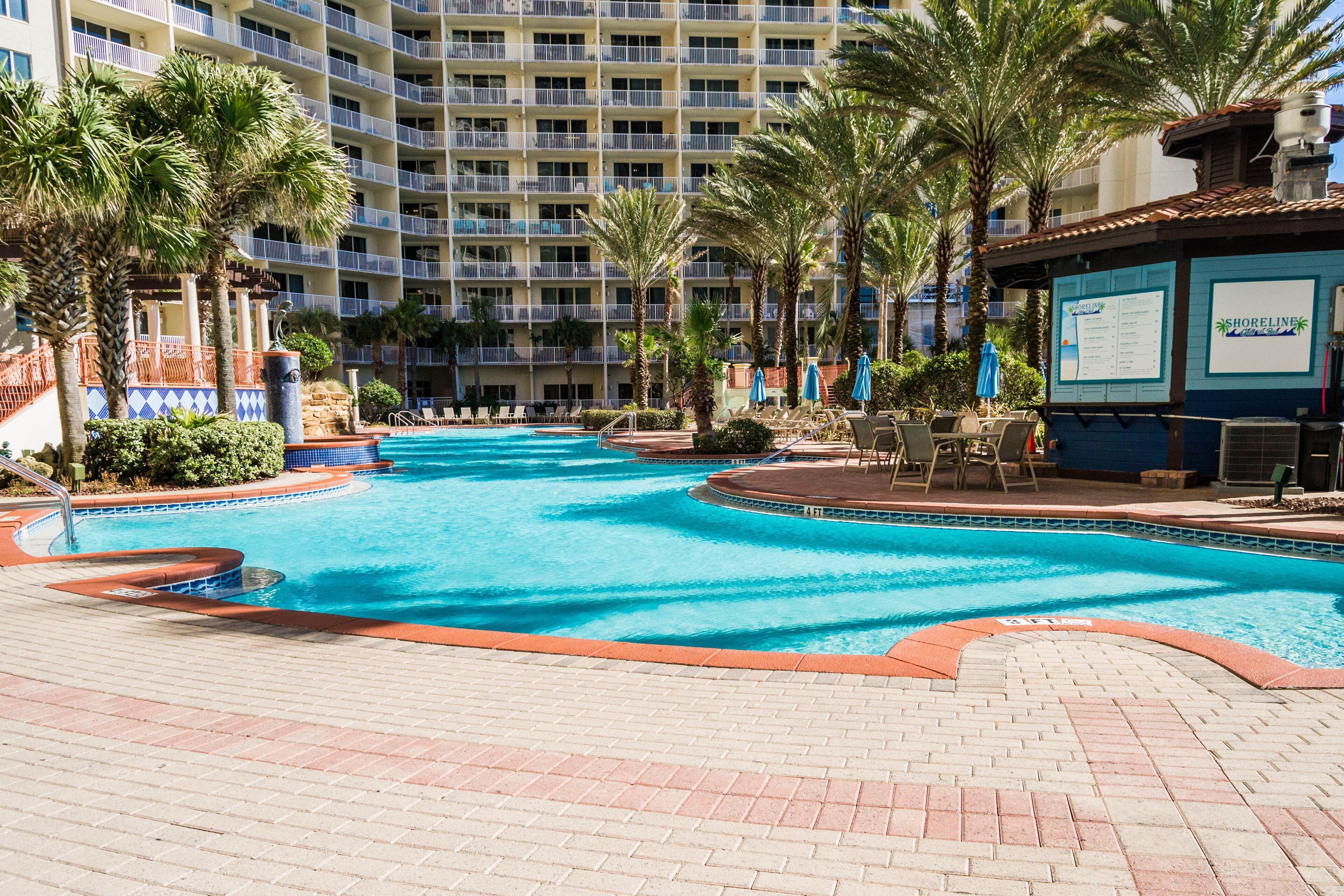 A hotel pool area with a clear blue pool, palm trees, and a building with balconies in the background.
