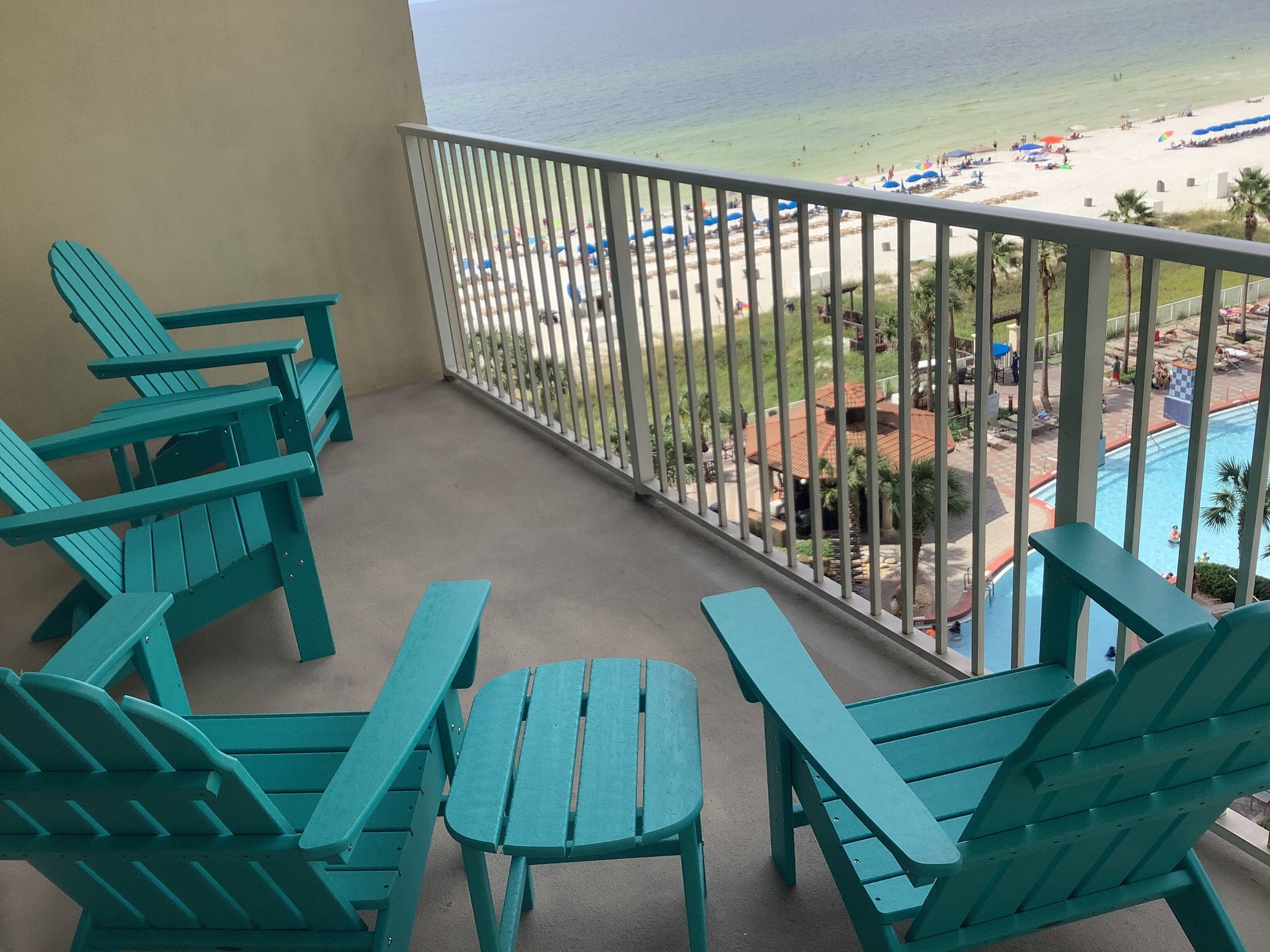 Un balcon avec des fauteuils Adirondack turquoise et une vue sur une plage parsemée de parasols et sur une piscine.