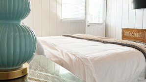 Une chambre à coucher comprenant un lit, une table de chevet avec un plateau en verre, une lampe de couleur turquoise, une tête de lit en bois, une fenêtre avec des stores et un téléviseur à écran plat fixé au mur.