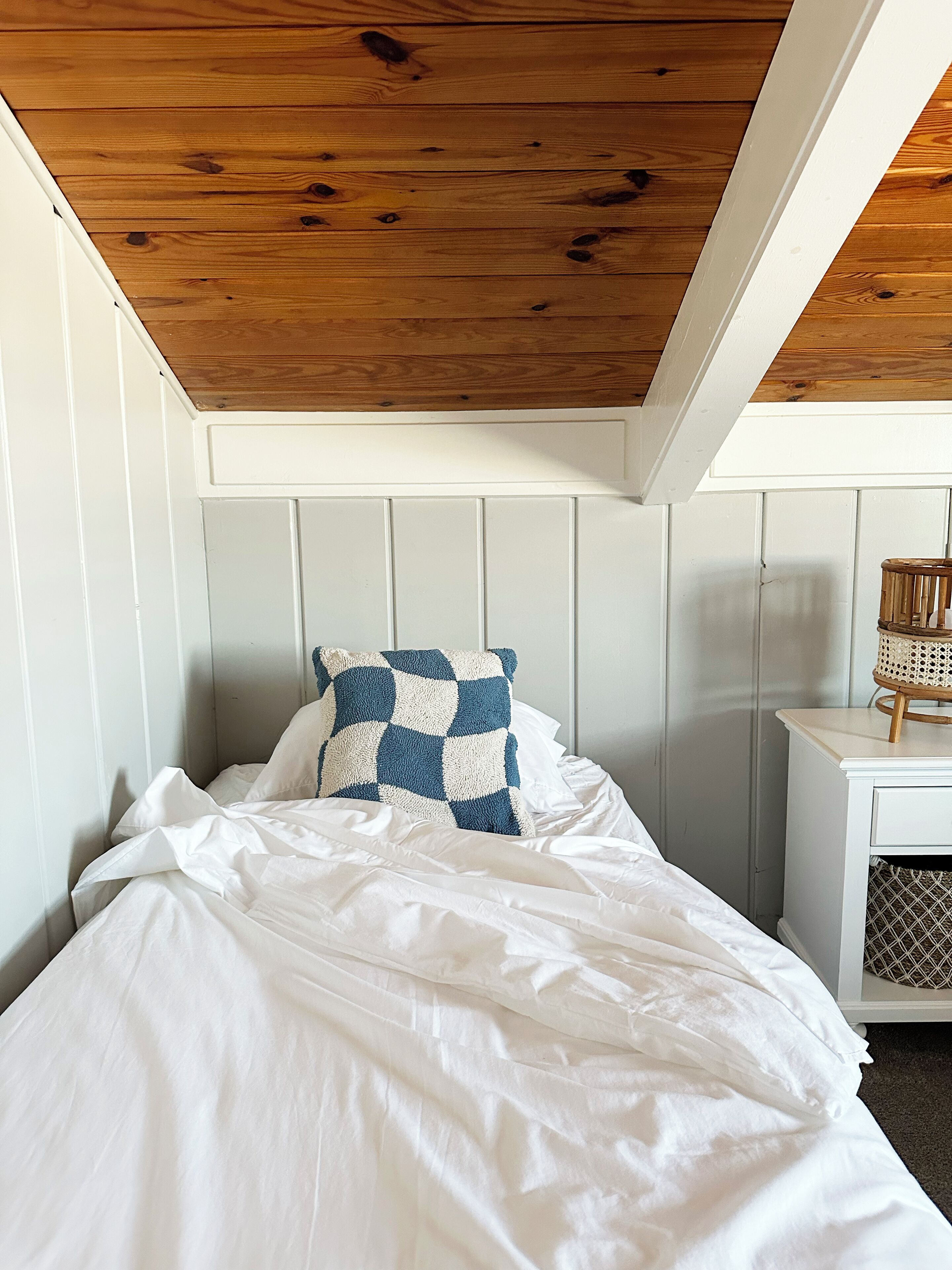 Ein Schlafzimmer mit geneigter Decke, weißen Wänden, einem Bett mit weißer Bettwäsche, einem blau-weißen Karokissen und einem kleinen Nachttisch mit einer Weidenkorlampen.
