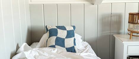 Ein Schlafzimmer mit geneigter Decke, weißen Wänden, einem Bett mit weißer Bettwäsche, einem blau-weißen Karokissen und einem kleinen Nachttisch mit einer Weidenkorlampen.