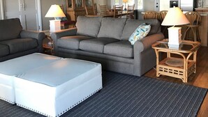 A cozy living room with a grey sofa, a white ottoman, and a dining area with wooden chairs and a table. - 709 Bluefish - Ocean Front (Saint Helena Island)