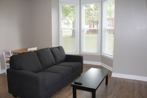 A living room with a dark grey sofa, a small black coffee table, and a wooden dining table with chairs. There are white-framed windows with blinds. - Drew's Den in Historic Downtown Pleasant Hill, MO (Pleasant Hill)