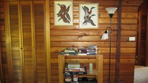 A cozy cabin interior with wooden walls, a bookshelf with books and board games, a lamp, and two framed pictures on the wall.