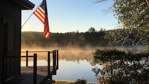A wooden dock extending over a calm lake with a misty backdrop, an American flag, and a forested hill in the distance.