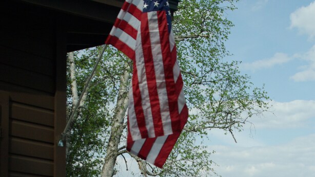 A wooden deck with a view of a lake, an American flag, and a red chair.