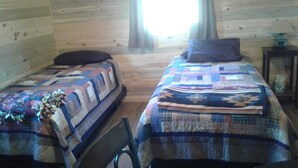 Dos camas con colchas, una pared de madera, una ventana con cortinas y una mesita con lámpara.