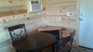 Interior de una cabaña de madera con una mesa redonda, dos sillas, un escritorio pequeño y una unidad de aire acondicionado montada en la pared.