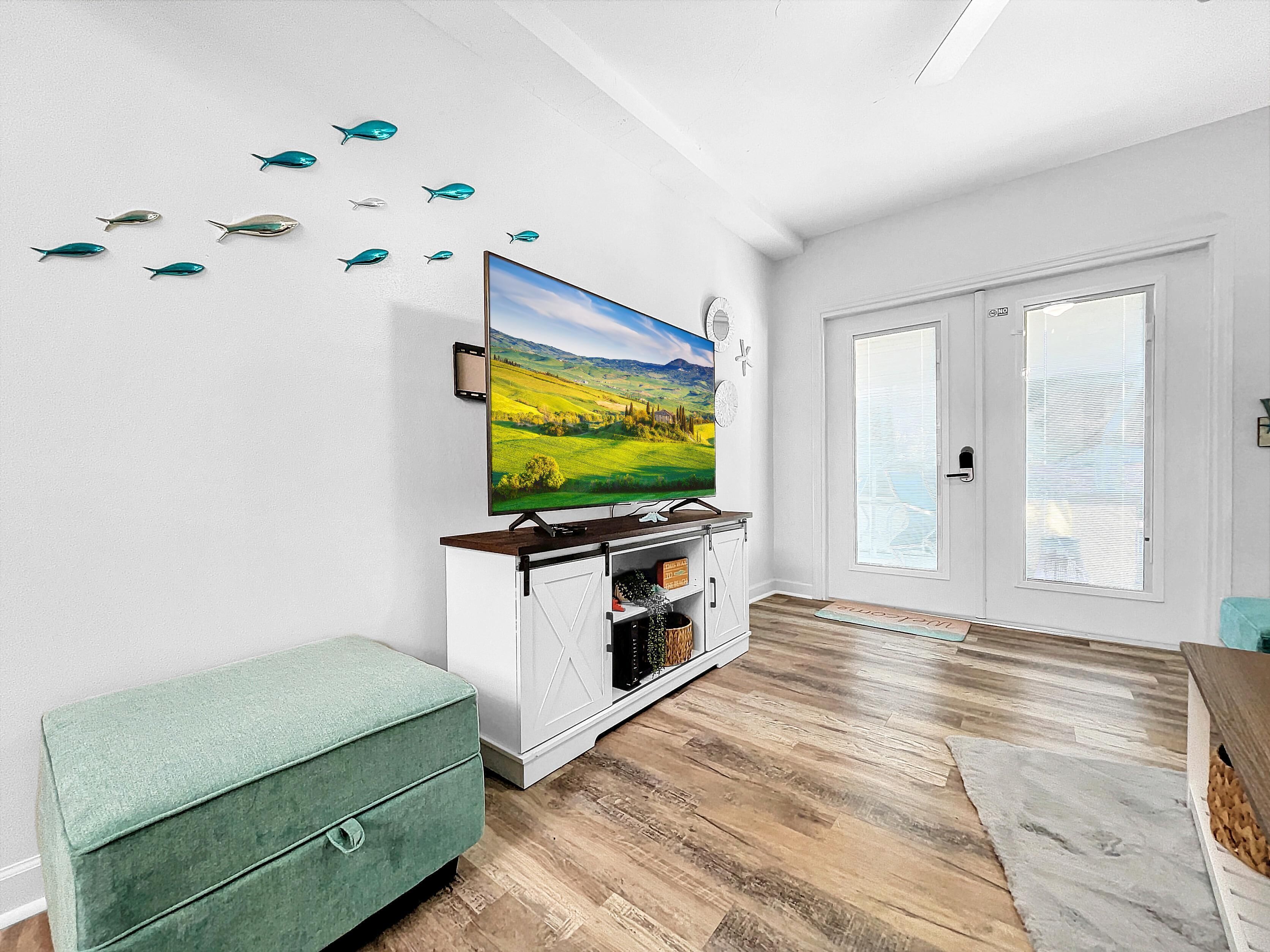 A living room with a large flat-screen TV mounted on the wall, a wooden cabinet below it, a green cushioned bench, and a wooden floor.