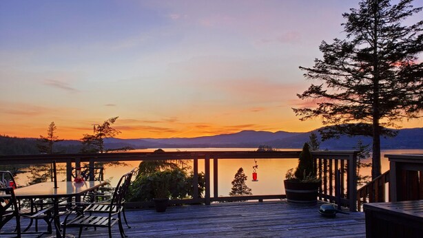 A wooden deck with outdoor seating, overlooking a calm lake with mountains in the background. - Architect Designed And Decorated Waterfront House, You Have The Whole House! (Sooke)