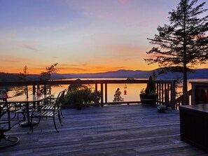 Una terraza de madera con asientos exteriores, con vista a un lago tranquilo y montañas al fondo.
