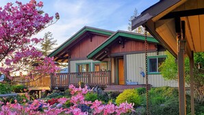 A wooden cabin with a balcony, surrounded by lush greenery and pink flowers. - Architect Designed And Decorated Waterfront House, You Have The Whole House! (Sooke)
