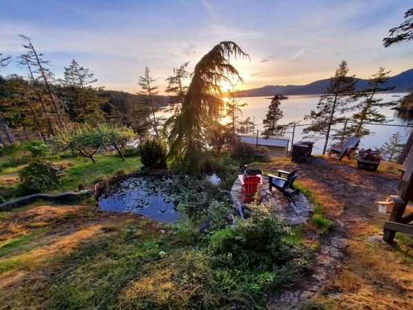 Un patio junto al lago con un brasero, sillas Adirondack y un depósito de leña de piedra.