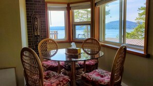 A dining area with a round glass table and wicker chairs, a brick wall, and large windows offering a view of a lake and mountains. - Architect Designed And Decorated Waterfront House, You Have The Whole House! (Sooke)