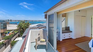 Un balcón con vistas a la playa, una puerta corrediza de vidrio que da a una habitación con una cama y una silla, y una pequeña mesa.