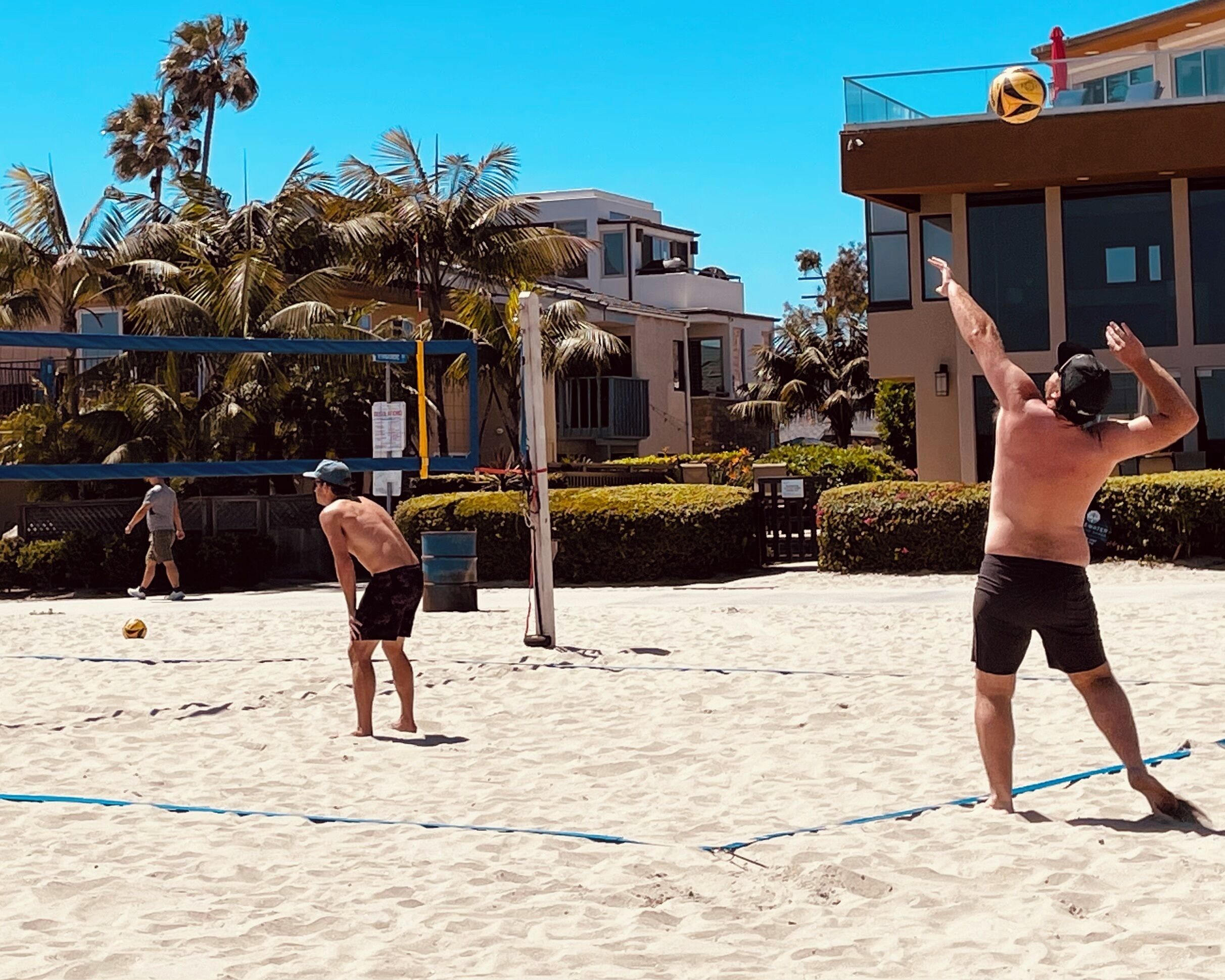 A beach volleyball game on a sandy court with palm trees and buildings in the background.