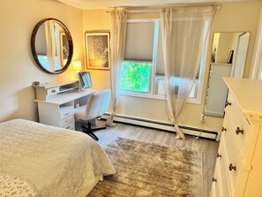 A bedroom with a bed, a desk, a chair, a dresser, a mirror, and a window with curtains.