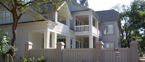 Une maison à deux étages avec un balcon au deuxième étage, une cour clôturée et un arbre à l’avant.
