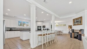 A spacious kitchen with white cabinetry, a central island with bar stools, and a dining area with a fireplace. - Ocean View, 3-Patio, 6BR, 6300sf Home w/Pool; 50 Steps to Beach, Coligny Circle! (Hilton Head Isalnd)