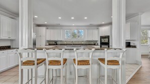 A spacious kitchen with white cabinetry, a central island with bar stools, and a large window providing natural light. - Ocean View, 3-Patio, 6BR, 6300sf Home w/Pool; 50 Steps to Beach, Coligny Circle! (Hilton Head Isalnd)