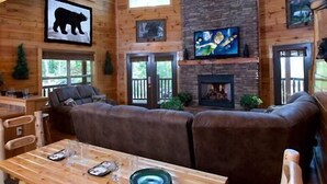 A cozy cabin interior with a fireplace, leather sofa, wooden dining table, and large windows. - A Bear's Kind of Fun in Pigeon Forge #unforgettable 4 bedroom w/ Games & Full Theater! (Sevierville)