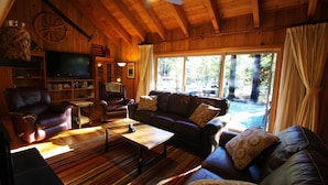 A cozy cabin interior with a wooden ceiling, a leather sofa, a television, and a large window overlooking a forest. - Charming Cabin Near Fort Rock Park and SHARC! Hot Tub! 8 SHARC passes included! (Bend)