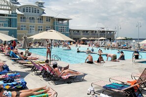 Un espace piscine doté de chaises longues et de parasols, entouré de bâtiments avec des balcons et d’une structure couverte.