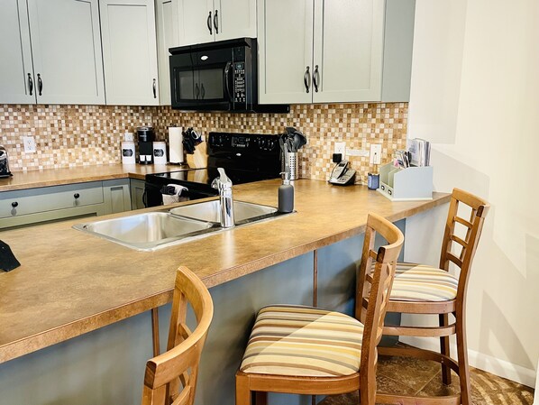 A kitchen with a double sink, a microwave, a coffee maker, and two wooden chairs with striped cushions.