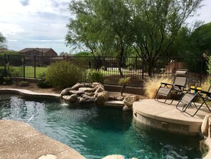 A swimming pool with a rock feature, surrounded by a fence and trees. - Brand New 51 Jet Spa, Heated Pool, Perfect Escape-Spring Training/Golf/Shopping (Peoria)