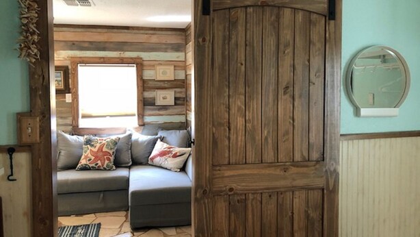A room with a sliding barn door, a sofa with decorative pillows, a window with a view, and a wall-mounted mirror. - The Shell Cabin is a place to relax and enjoy the beach! (Port Aransas)