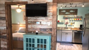 A rustic room with a bed, a small cabinet, a TV, and a kitchen area with a refrigerator and cabinets. - The Shell Cabin is a place to relax and enjoy the beach! (Port Aransas)