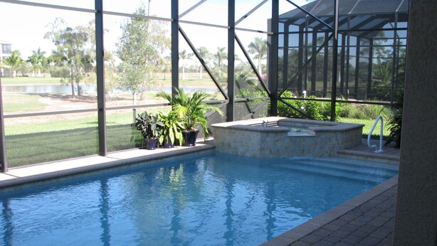 An indoor swimming pool with a clear view of the surrounding landscape, including a grassy area and trees. - Model home in Lely Resort Naples Florida (Naples)