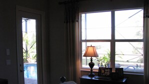 A hotel room with a bed, a desk with a lamp, a window with a view of a swimming pool and outdoor plants. - Model home in Lely Resort Naples Florida (Naples)