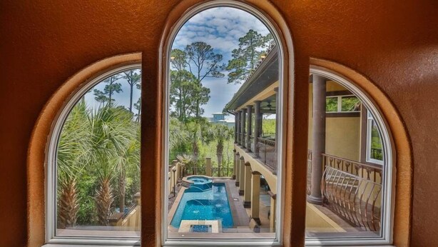 A view through a window of a pool area with a pool, palm trees, and a building with columns and a balcony. - Casa Bahari W/Private Pool 5-Bed/Pool/Spa/Lux Amenities (Destin)