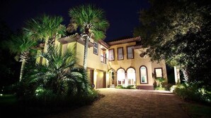 A well-lit building with a paved entrance, surrounded by palm trees and greenery. - Casa Bahari W/Private Pool 5-Bed/Pool/Spa/Lux Amenities (Destin)
